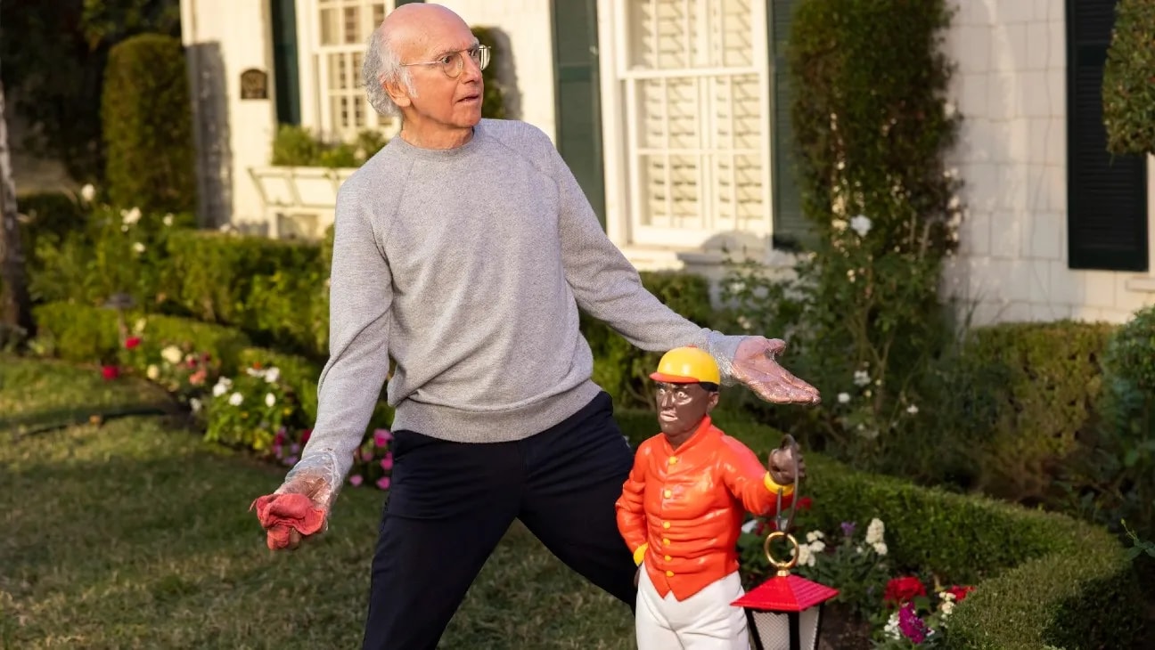 Larry David beside a small jockey figure in a garden